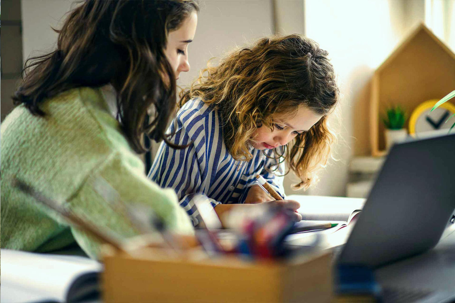 Mutter sitzt neben ihrem Kind und hilft beim Schreiben und Lernen am Tisch mit Laptop – Sinnbild für Strahlenquellen im Alltag.