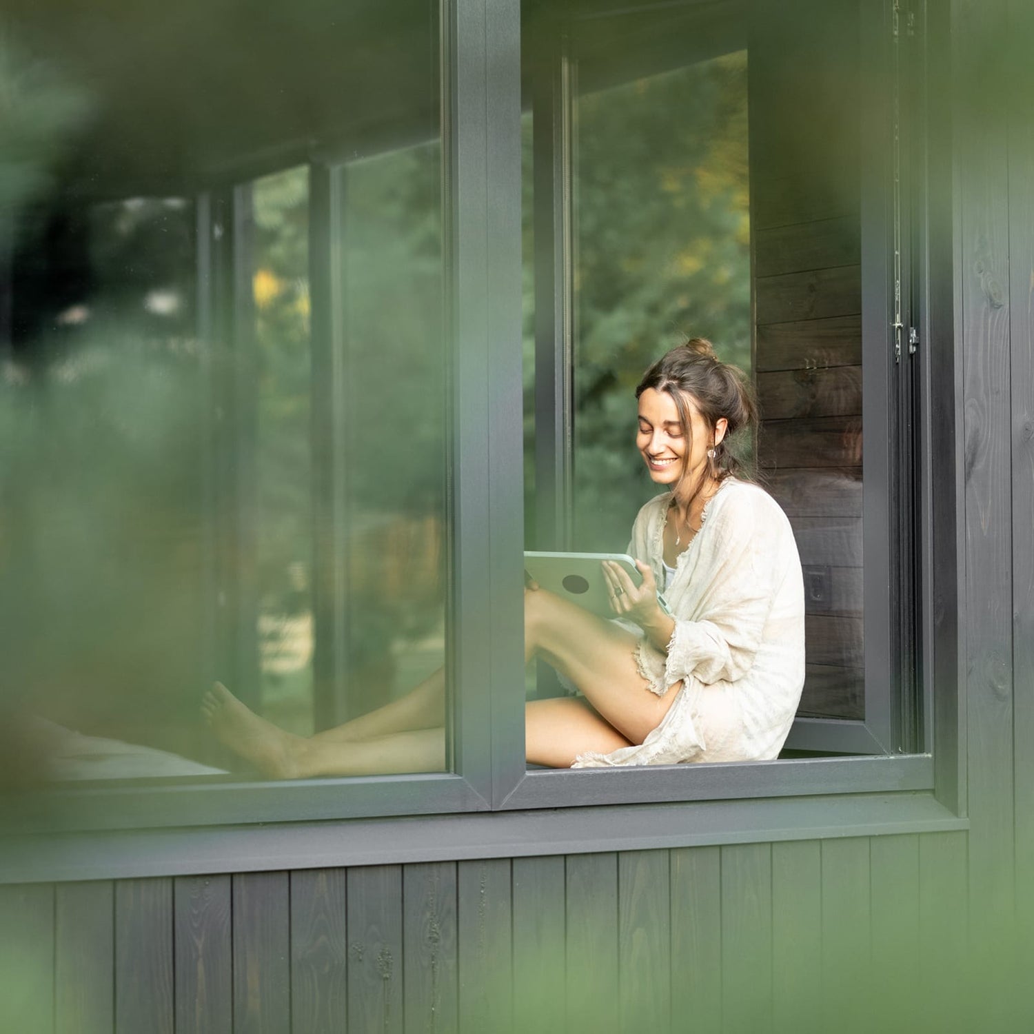 Junge Frau sitzt entspannt am Fenster eines modernen Hauses und liest auf einem Tablet – Sinnbild für Ruhe, Achtsamkeit und bewussten Umgang mit Technologie.